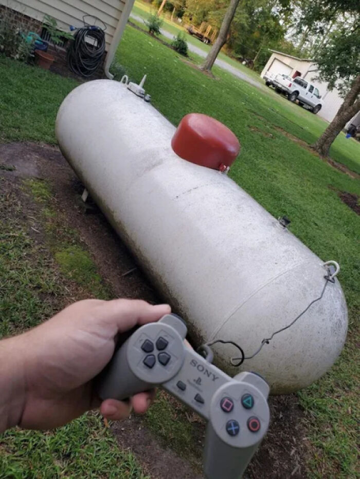 Person holding a PlayStation controller pointed at a large propane tank outside, creating a humorous visual.