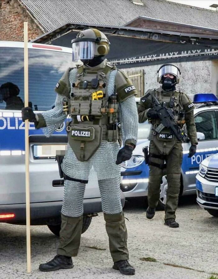 Two individuals in police gear, one wearing chainmail and holding a stick, as if blending with law enforcement.