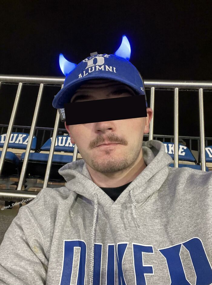 Person wearing Duke hoodie and hat with glowing horns, sitting in stadium, exemplifying cheat the system behavior.