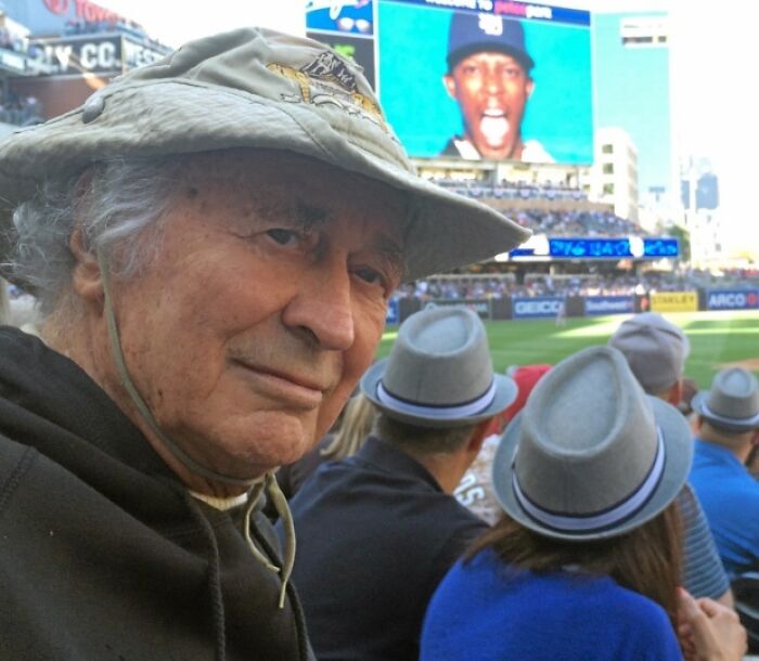 Elderly man in a sunhat at a sports game, blending into the crowd; screen in the background showing a player's photo.