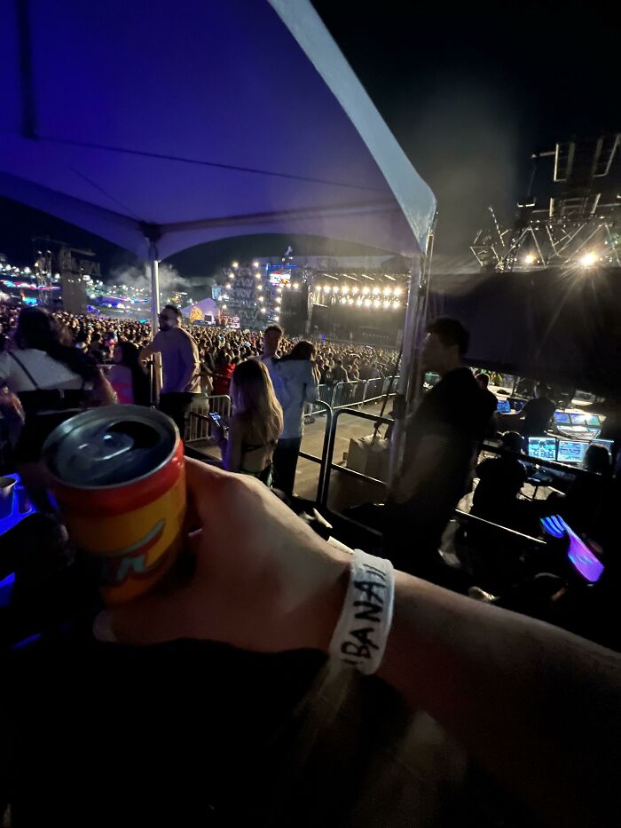 Hand holding a drink with a "BANANA" wristband, standing by a crowded concert stage, blending in with the scene.