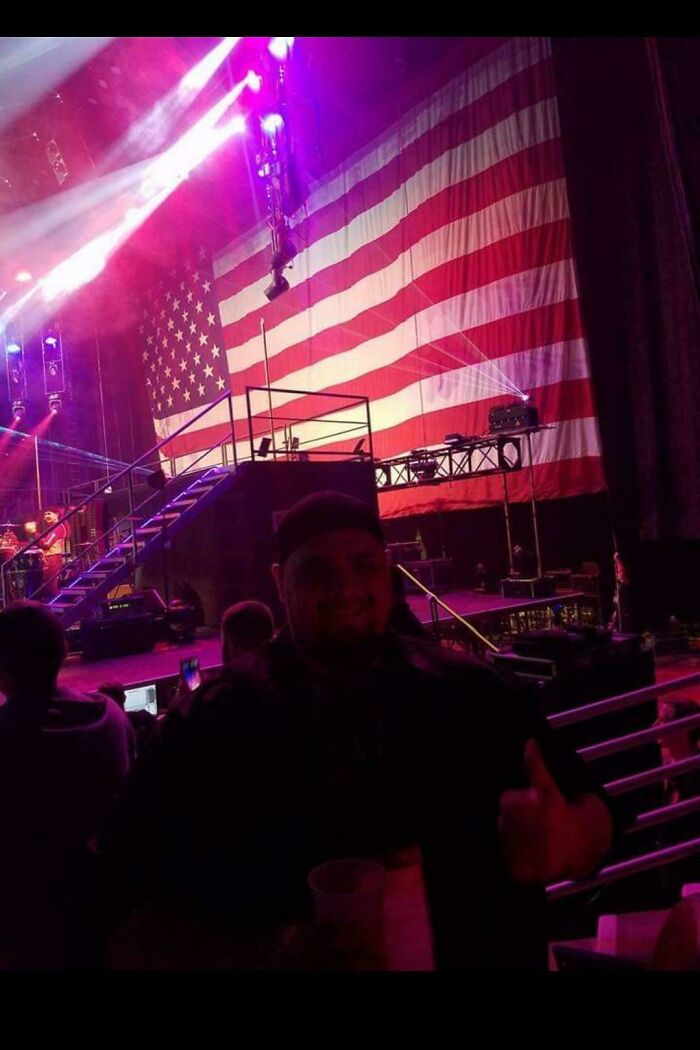 Person at music event, giving thumbs up in front of large American flag, blending in to cheat the system.