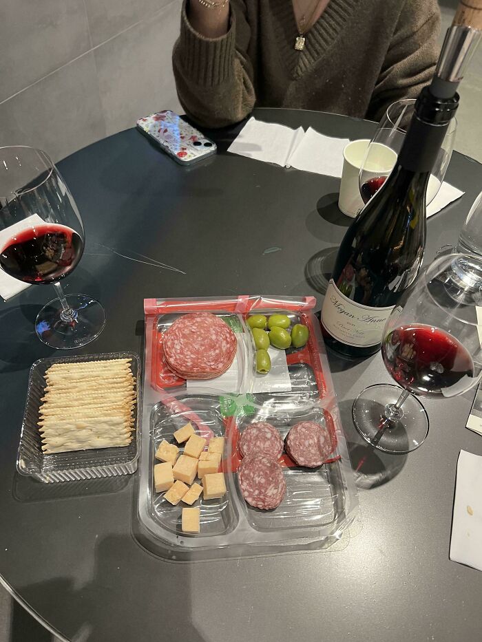 Charcuterie board served in a plastic snack tray with crackers and glasses of red wine at a restaurant table.