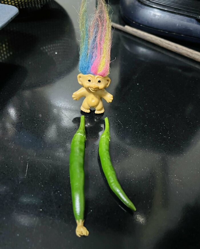 Troll doll with colorful hair next to two green peppers on a black surface, highlighting hilarious harvesting fails.
