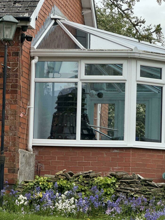 British humor captured with a Dalek inside a conservatory, visible through a window, in a classic UK garden setting.