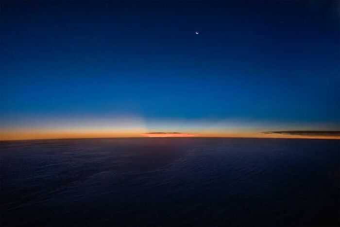 A breathtaking aerial photo showing a twilight sky with a crescent moon above a dark horizon taken by a pilot.
