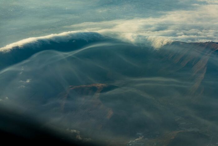 A breathtaking aerial photo showing mist and clouds rolling over a mountainous landscape captured by a pilot in flight.