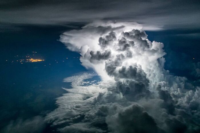 Aerial photo of dramatic illuminated storm clouds at night with distant city lights visible on the horizon.