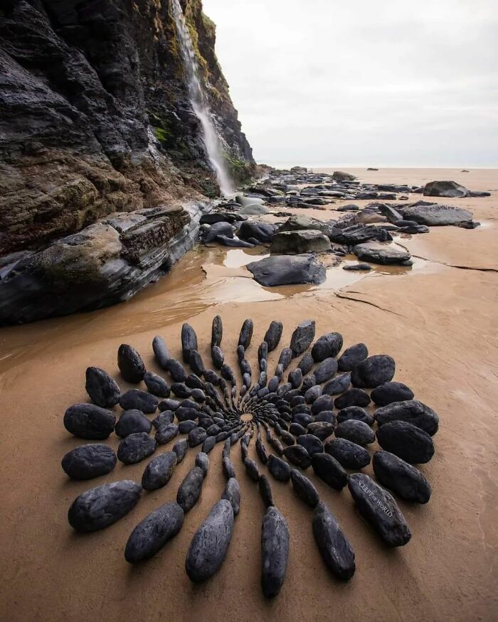 Spiral land art made with natural stones arranged on a sandy beach near rocky cliffs in an outdoor setting.