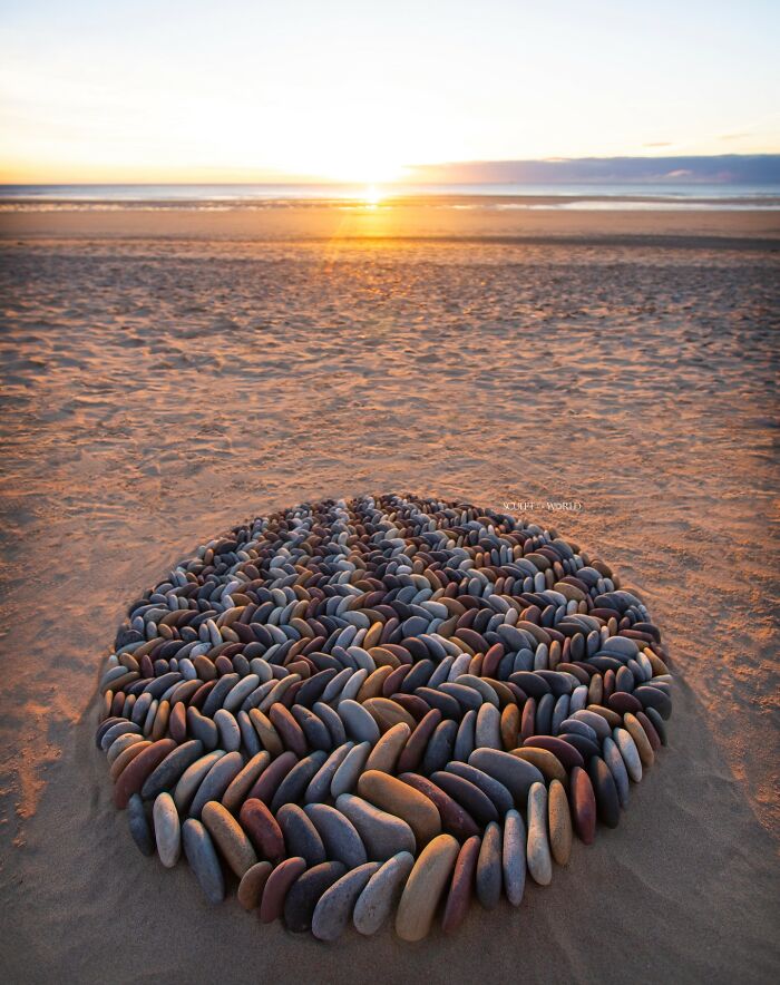 Intricate land art piece made with natural stones arranged in a circular pattern on a sandy beach at sunset.