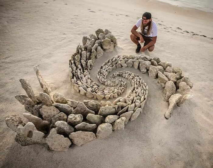 Artist creating a patience-requiring land art piece with natural stones arranged in an intricate spiral pattern on sand.