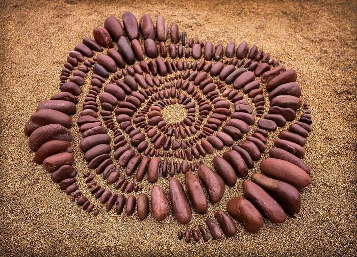 Spiral land art piece made with natural materials featuring smooth brown stones arranged on sandy ground.