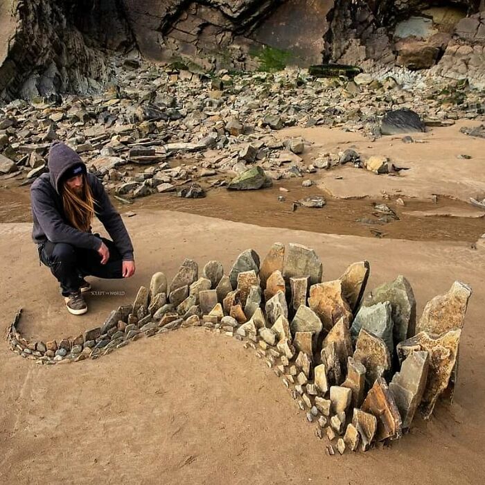 Artist creating patience-requiring land art piece with natural stones arranged on sandy ground near rocky cliffs.