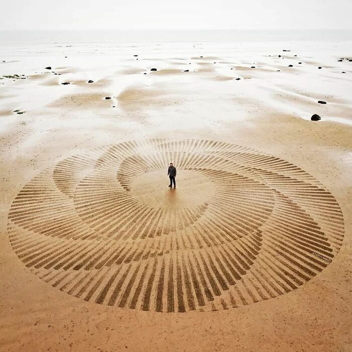 Large land art piece made with natural materials on a beach featuring intricate spiral patterns and a person in the center.