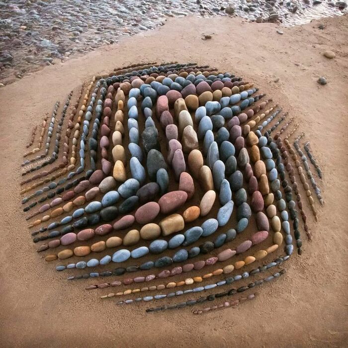 Land art piece with colorful natural stones arranged in layered, flowing patterns on sandy ground.