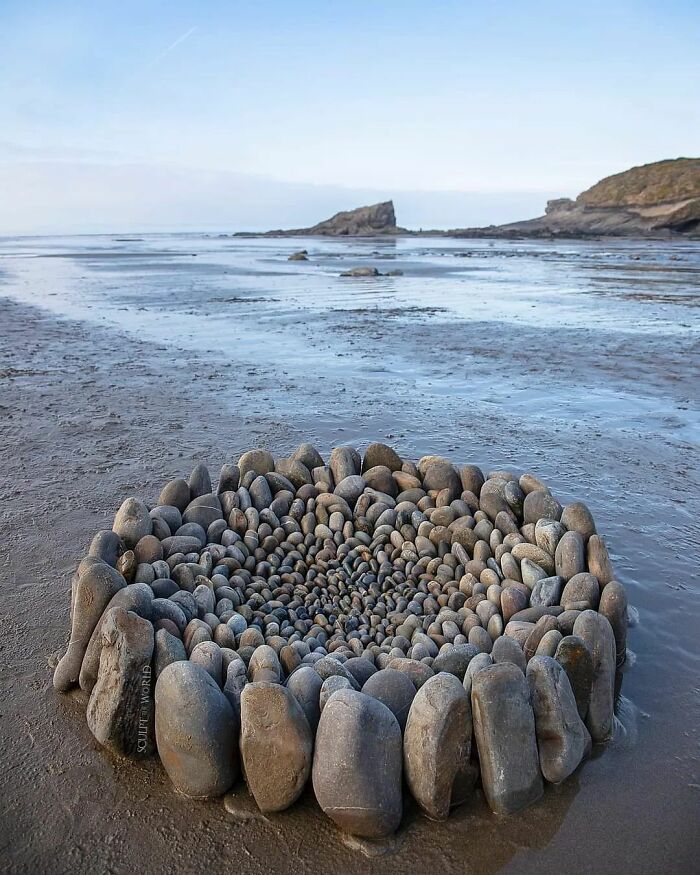 Intricate land art piece made of natural stones arranged in a spiral pattern on a beach shore.