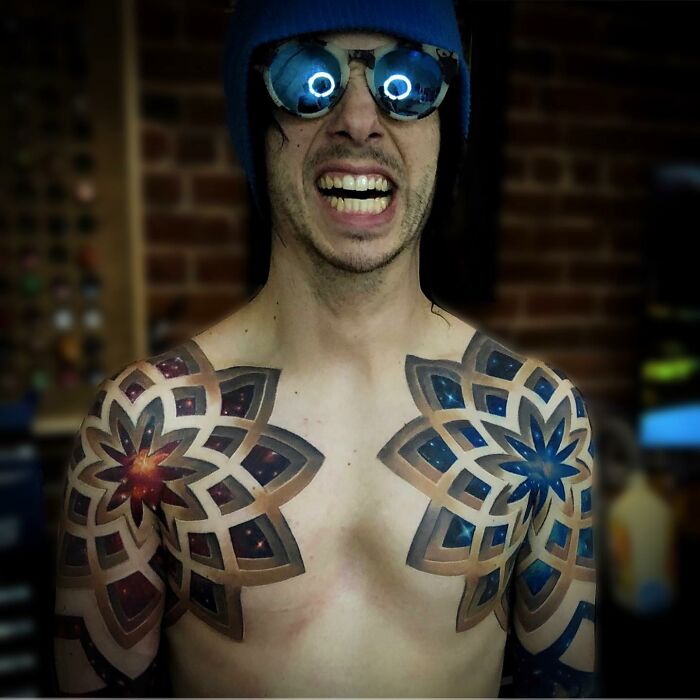 A person with intricate mandala tattoos on shoulders, wearing a blue hat and reflective sunglasses, smiling broadly.
