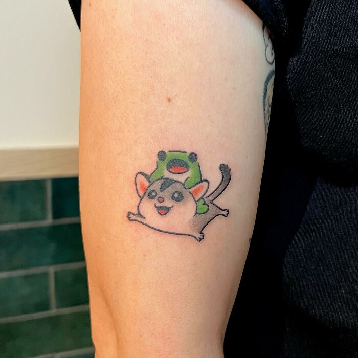Charming tattoo by Buoy featuring a cute animal character with a frog hat on an arm.