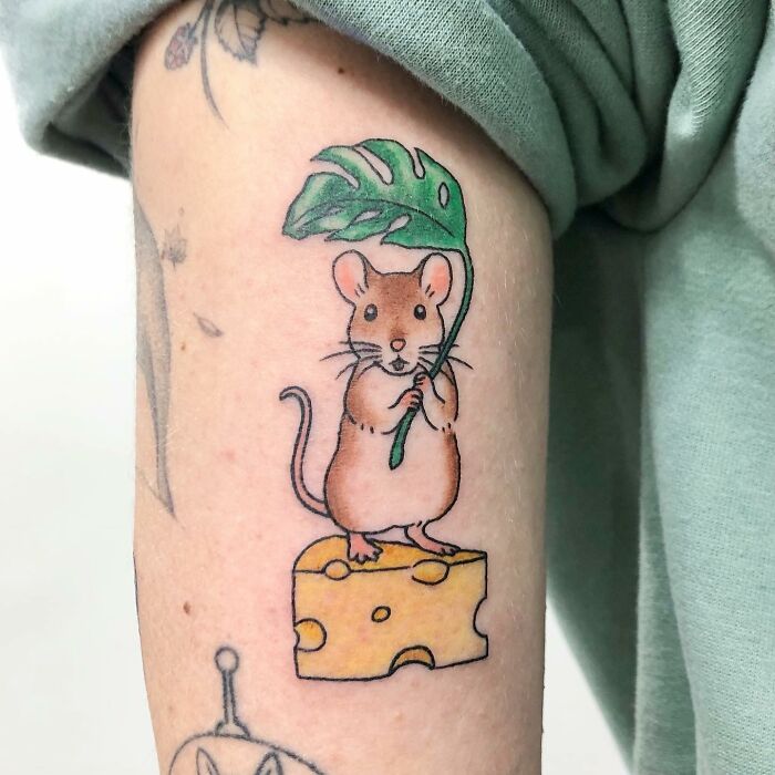 A charming tattoo of a mouse on cheese with a leaf umbrella, showcasing Buoy's artistic style.