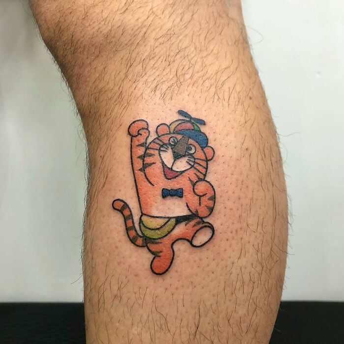 Charming tiger tattoo by Buoy, featuring a cartoon design with a blue hat and bow tie on a person's leg.