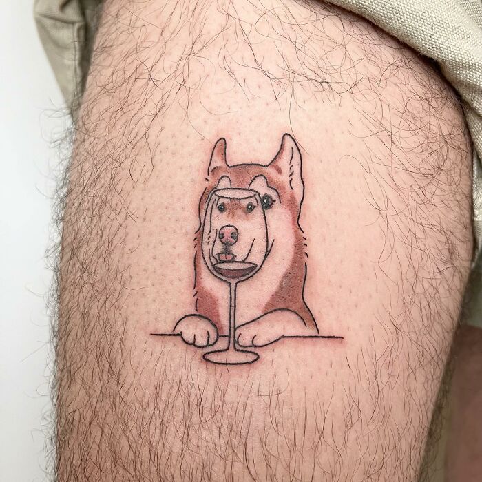 A charming tattoo by Buoy of a dog looking through a wine glass, inked on a person's leg.