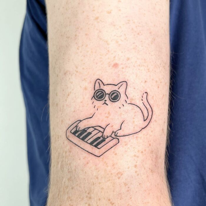 Tattoo of a charming cat wearing sunglasses and playing a keyboard on an arm.