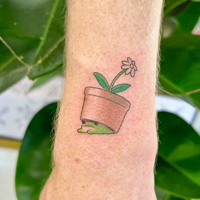Adorable tattoo of a flower in a pot with a cute face drawn on a forearm, by artist Buoy.