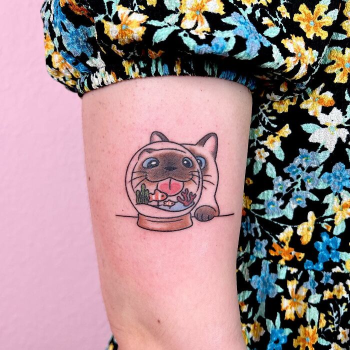 Charming tattoo of a cat peeking behind a fishbowl on an arm with a floral sleeve.