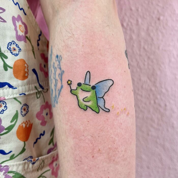 Charming tattoo of a whimsical green creature with wings on an arm, surrounded by colorful floral fabric.