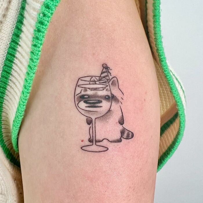 Charming tattoo by Buoy of a cute raccoon peeking through a martini glass, wearing a party hat.