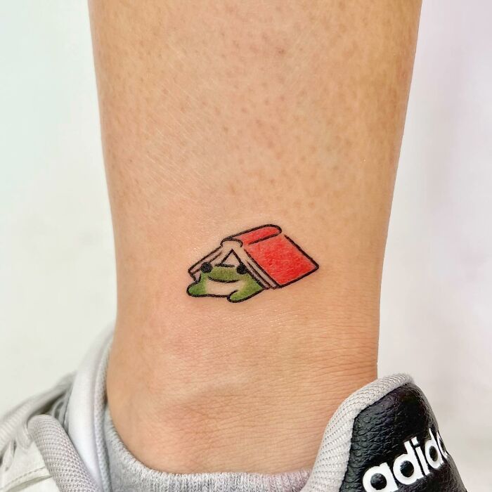 Charming tattoo of a small frog under a red book on an ankle, near an Adidas shoe.