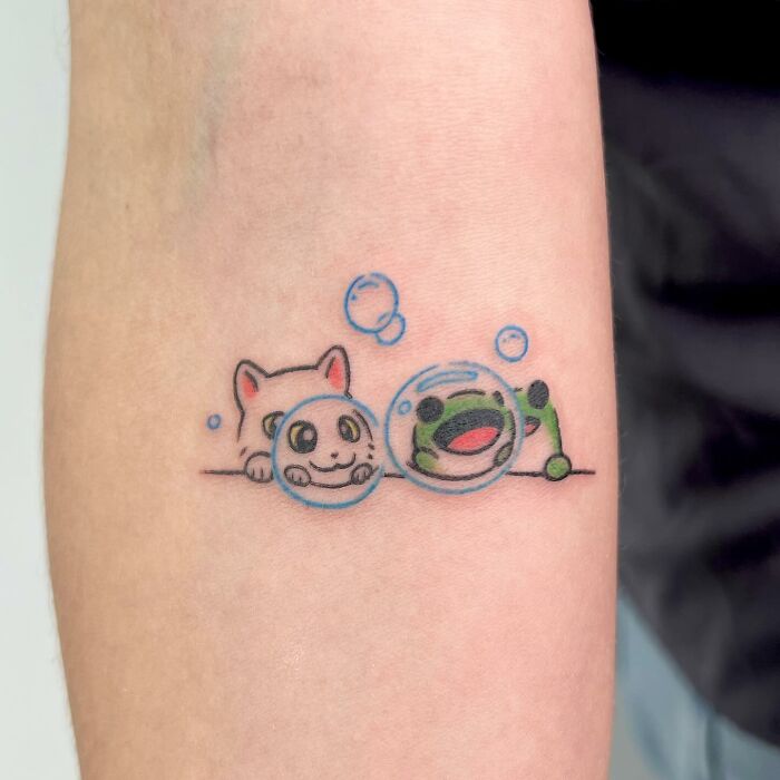 Charming tattoo by Buoy featuring a playful cat and frog with bubbles on a forearm.