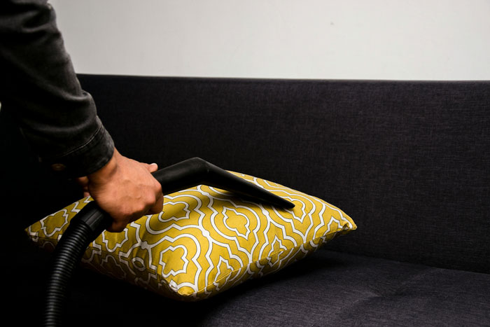 Person using vacuum nozzle to remove pet hair from a patterned pillow on a dark sofa. - 3