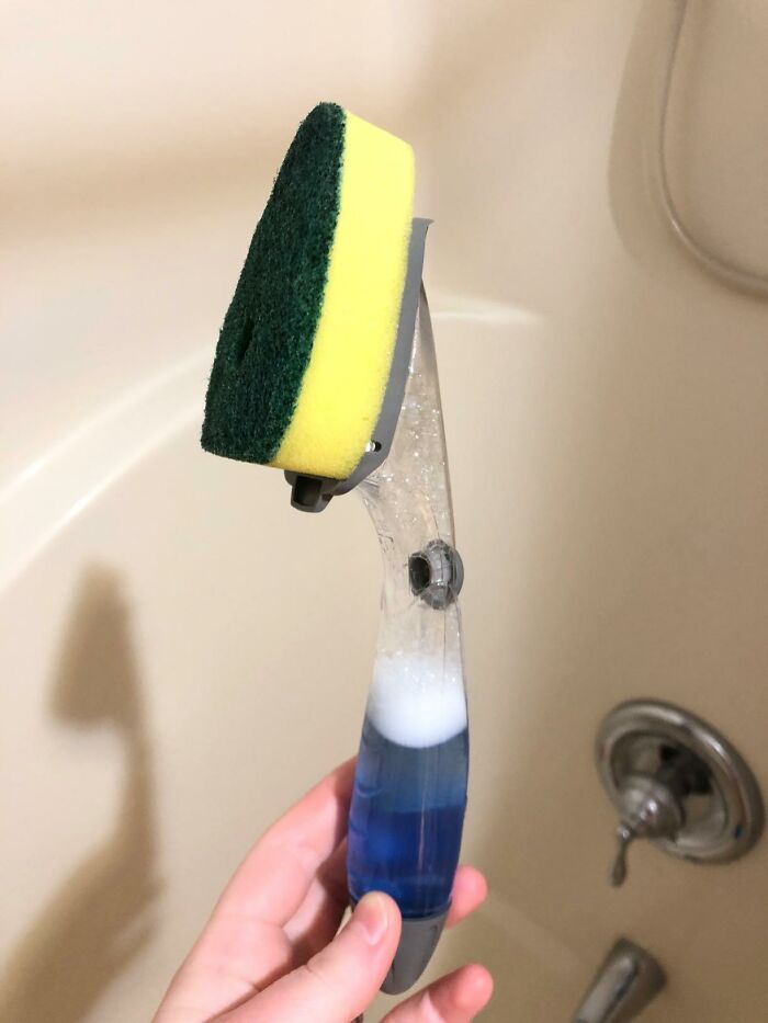 Hand holding a cleaning scrub brush with a sponge head and blue liquid soap, ideal for frantic panic cleaning before guests arrive.