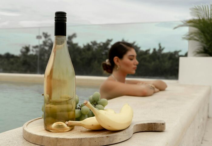 Bottle of white wine with bananas and grapes on a tray by the pool at a luxury resort with a woman relaxing nearby.