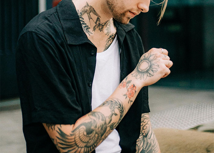 Man with tattoos on neck and arms wearing a black shirt, seated outdoors. - 1