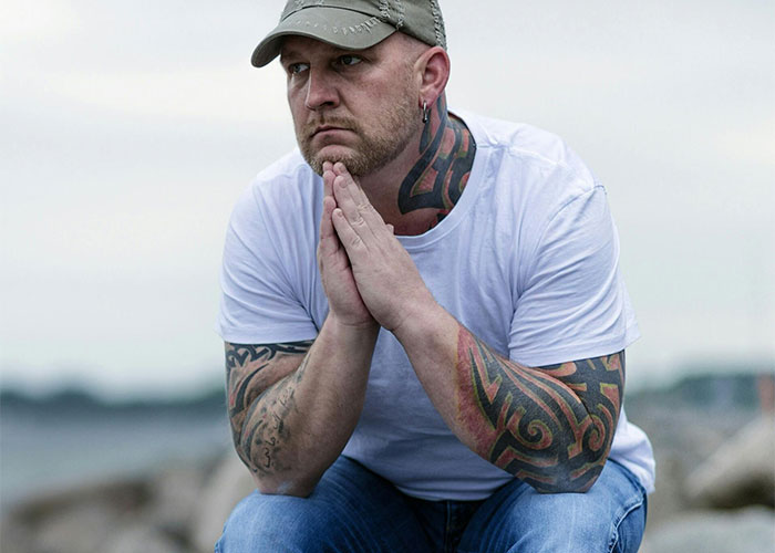 Man with tattoos sits pensively by the sea, wearing a white shirt and cap. - 13