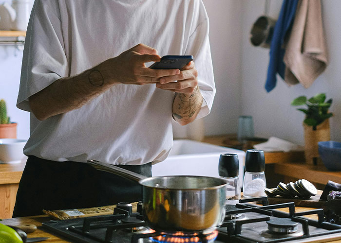 Person cooking in a kitchen, capturing a moment on their phone, illustrating shocked reactions to how others live.