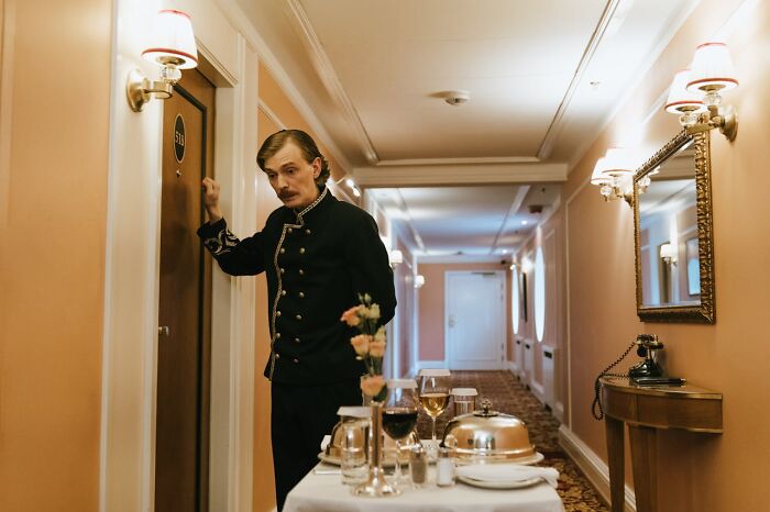 Luxury resort employee in uniform knocking on a guest room door with a cart of food service in a hotel hallway.