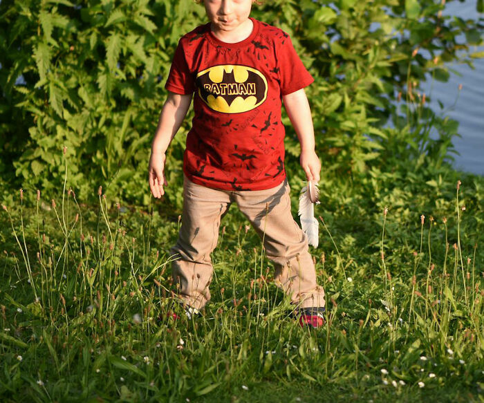 A child outdoors wearing a playful red "Ratman" shirt, standing on grass with greenery in the background.