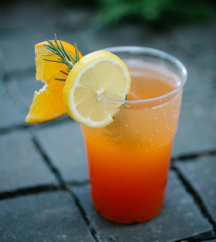 Orange and lemon cocktail in a clear cup on stone tiles, garnished with rosemary. - 3