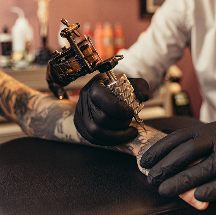 Tattoo artist working on arm, black gloves on, using tattoo machine, focused on intricate memorial tattoo design. Tattoo artist working on arm, black gloves on, using tattoo machine, focused on intricate memorial tattoo design.
