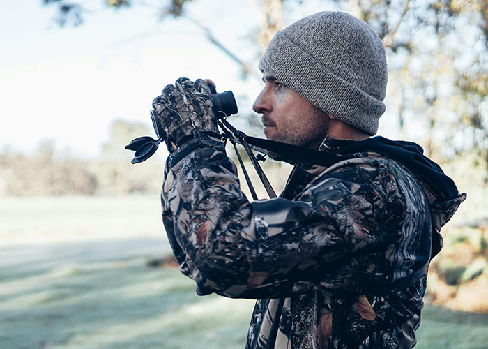 Man in camouflage jacket and beanie looking through binoculars outdoors, illustrating wildest things rich can purchase.