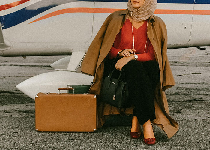 Woman in stylish coat and scarf seated by private plane with luxury suitcase and handbag, showcasing wildest things rich can purchase.