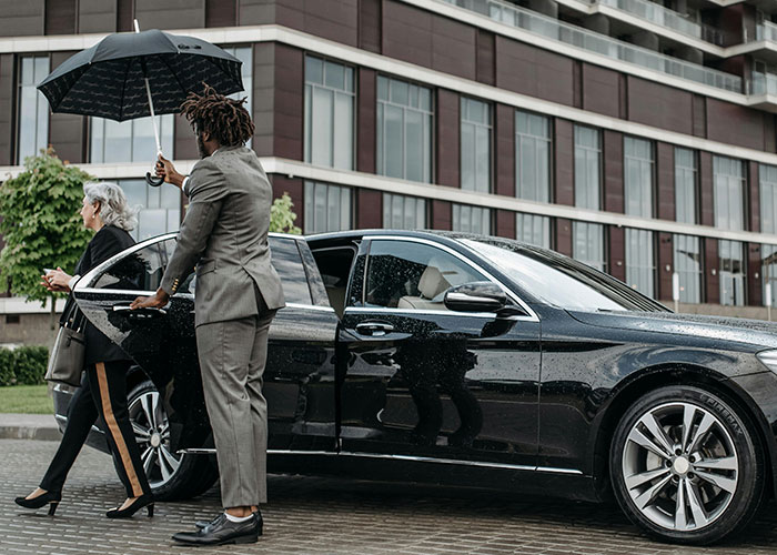 A personal chauffeur holding an umbrella for a woman exiting a luxury black car, showcasing wildest things rich can purchase.