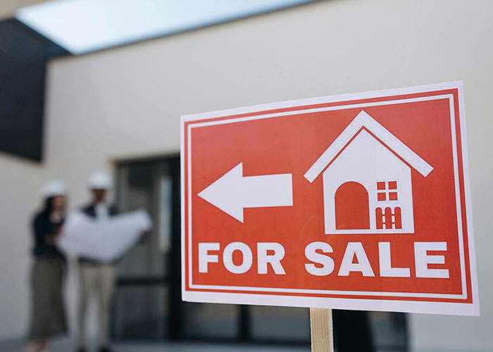 Real estate for sale sign in front of a house with blurred people reviewing property plans, luxury purchases concept.
