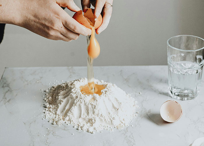 Hands cracking an egg into a flour mound on marble surface, illustrating unfiltered food opinions and cooking process.