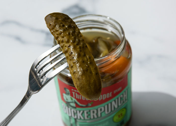 Pickle speared by a fork above an open jar of crunchy vinegar pickles on a marble surface.