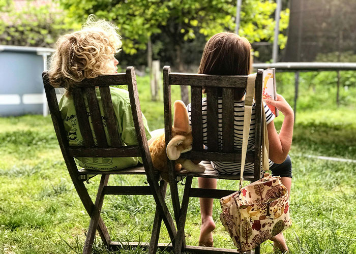 Two children sitting on wooden chairs outdoors with a stuffed animal, illustrating strange childhood realities.