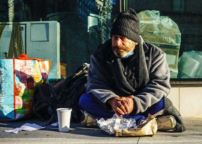 Homeless man sitting on sidewalk with bags and food, representing strange childhood realities people later learned were not normal.